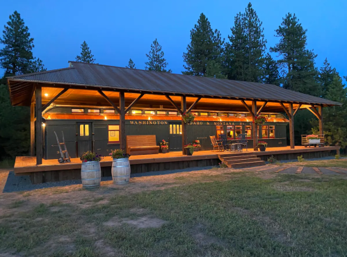 A rustic building with a covered porch, surrounded by trees, illuminated at dusk. Wooden barrels and seating are visible.